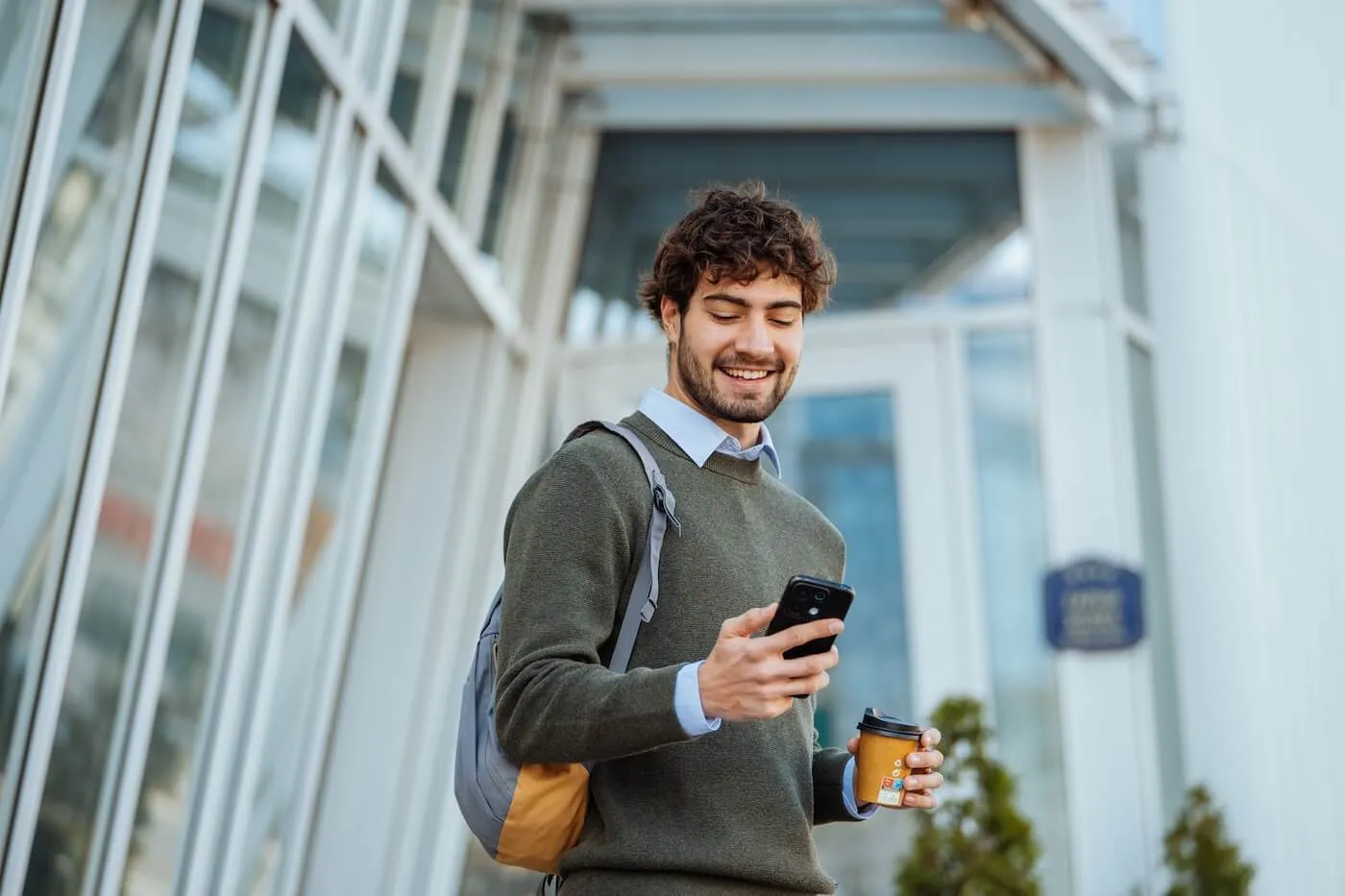 Smiling young man with a backpack looks at a smartphone while holding a coffee cup outside a glass office building