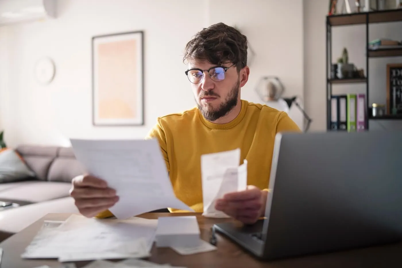 Focused young man seated at a table in a home interior reviews printed documents while holding a receipt, with an open laptop on the table and additional papers spread out, and shelving, framed artwork, and a sofa visible in the background.
