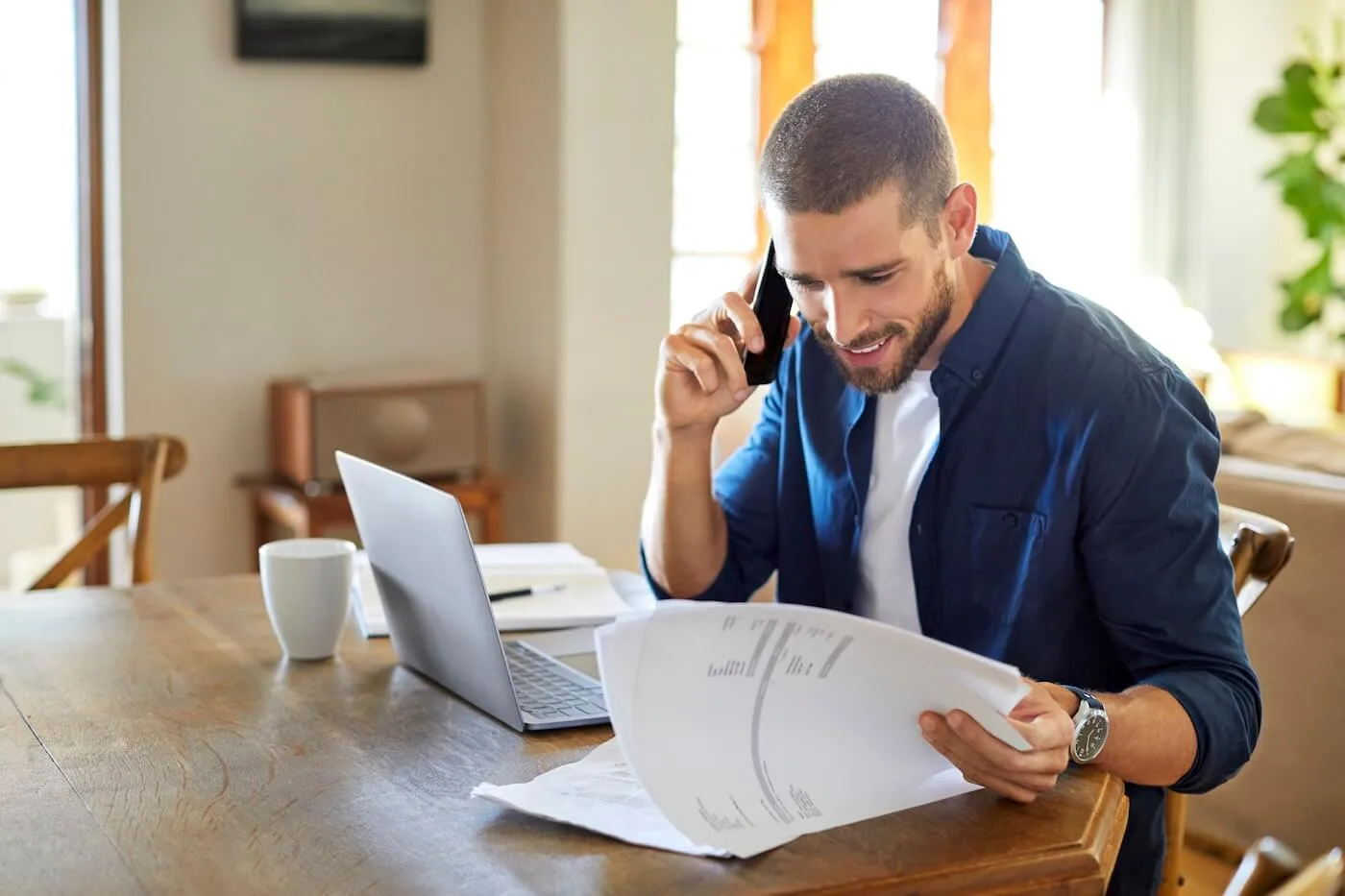 Young man reviews financial documents while speaking on a phone at a table with a laptop and coffee cup