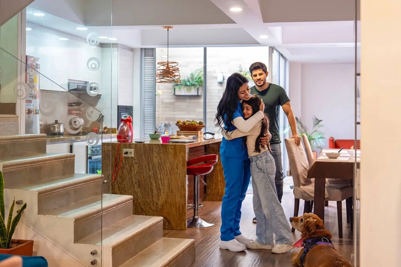 A family of three stands together in a modern kitchen and dining area, with the woman in a nurse uniform embracing a girl while the man stands close by. A dog lies on the floor near the group.