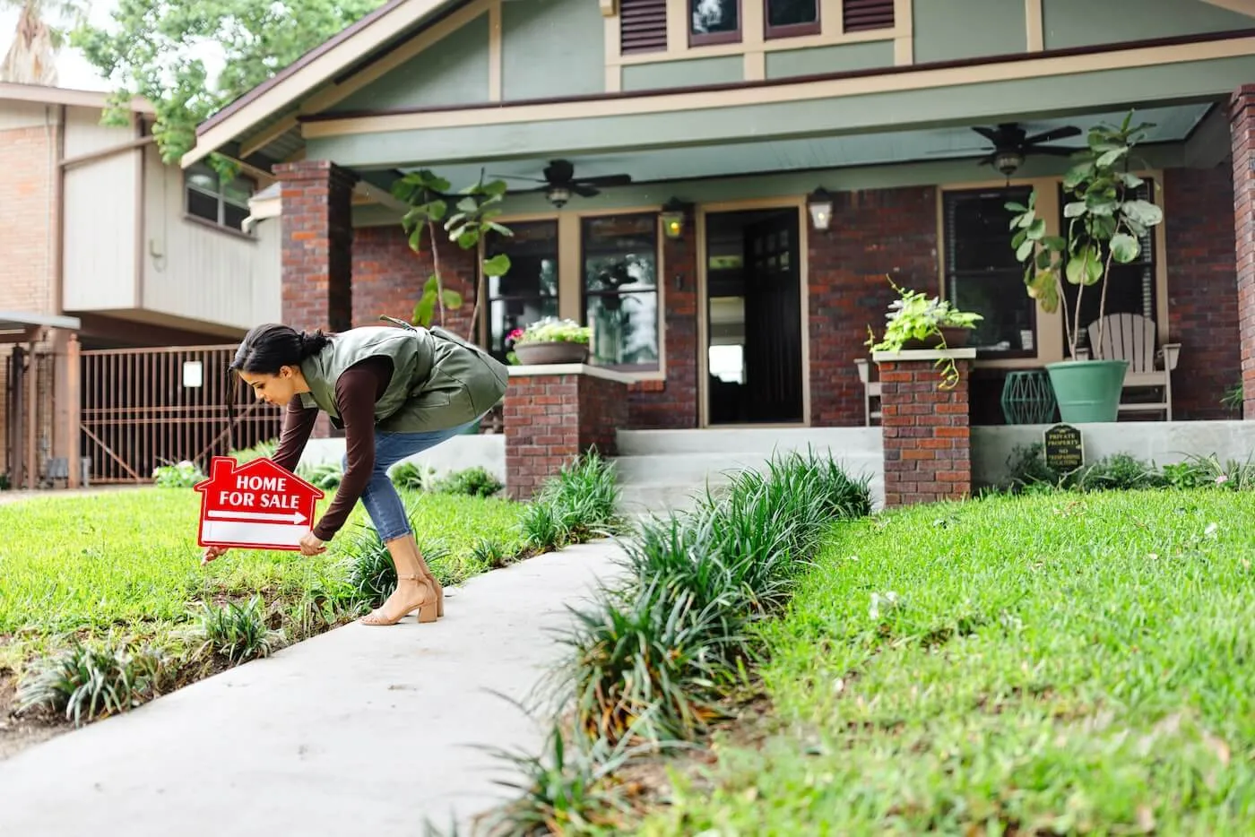 A real estate agent places a red “Home for Sale” sign into the front yard of a brick house, beside a curved walkway with plants and a covered porch in the background.