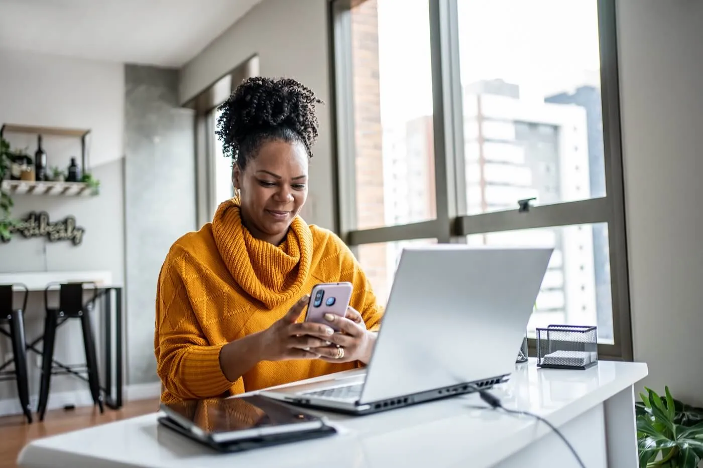 Smiling woman sits at a desk using a smartphone, with an open laptop, tablet, and office supplies on the desk near a large window.