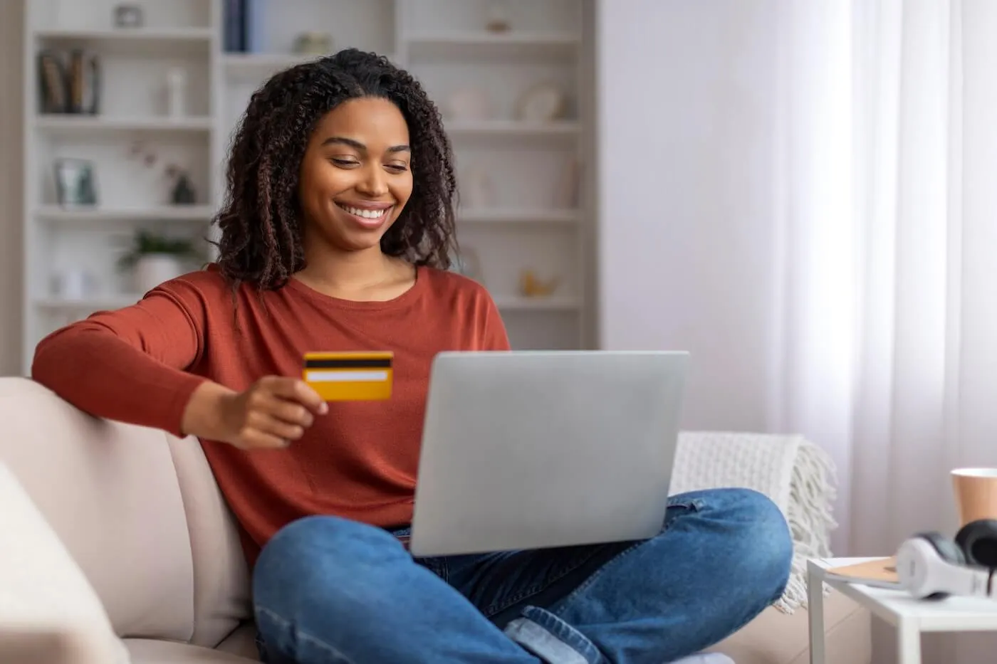 Smiling young woman uses her credit card while sitting on a couch with a laptop