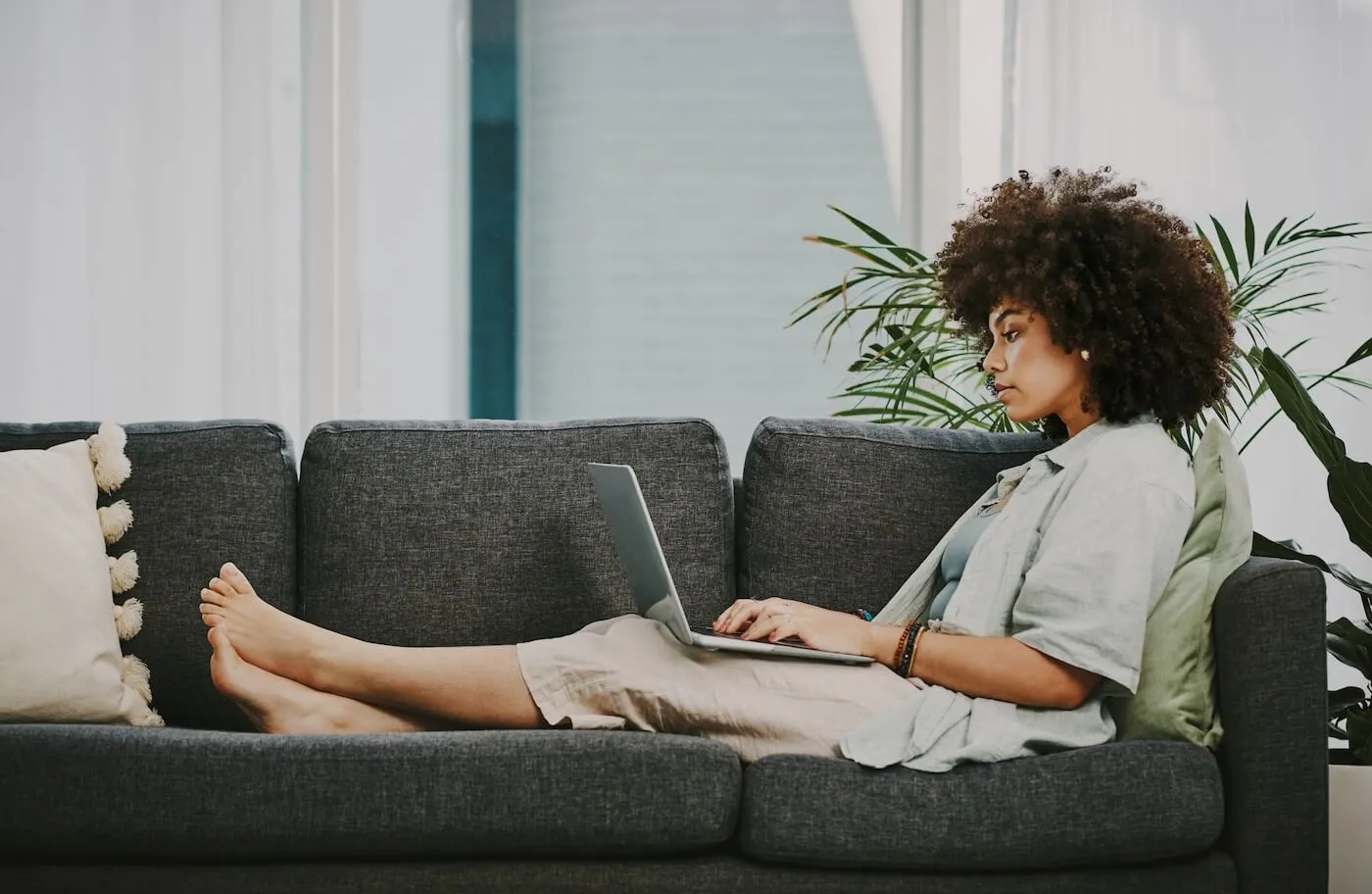 Focused woman reclines on a sofa while using a laptop, with legs stretched out, a pillow and plants nearby, in a bright living room setting.