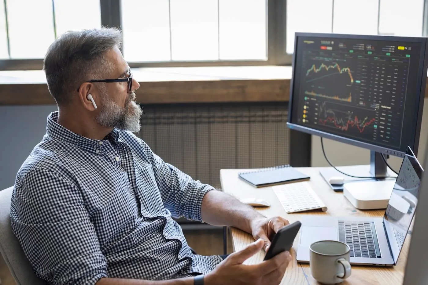 Focused mature man with earbuds sitting at a desk holding a smartphone while looking toward a monitor displaying stock charts and financial data, with a laptop, keyboard, notebook, and coffee mug nearby