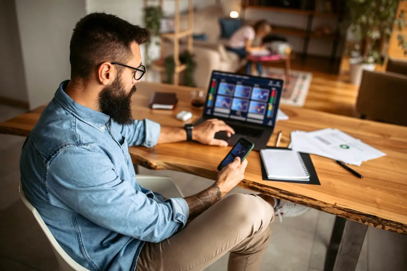 Focused man sitting at a wooden table using a smartphone while a laptop displays multiple charts; notebooks, papers, and a pen are spread out nearby in a home workspace.