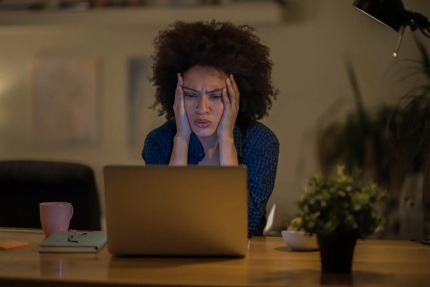 Concerned woman looking at the laptop monitor in a dimly lit room, with both hands placed on the sides of her face. A mug, a notebook, and a small potted plant sit on the desk, while a desk lamp casts warm light and a softly blurred interior is visible in the background.