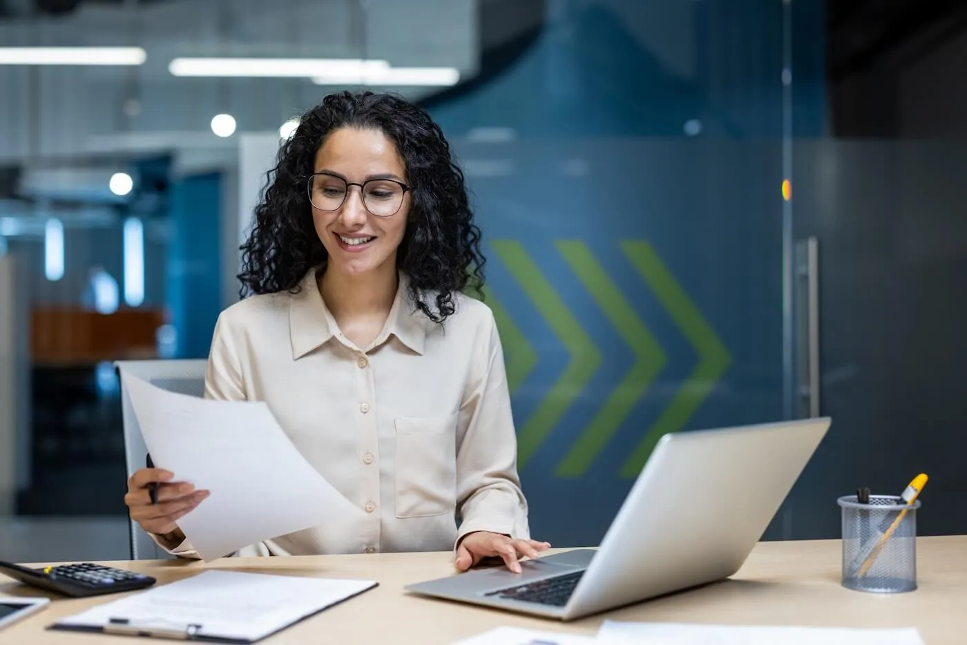 Smiling businesswoman sitting at an office desk holding a document while working on a laptop, with files, a calculator, and papers spread out on the table.