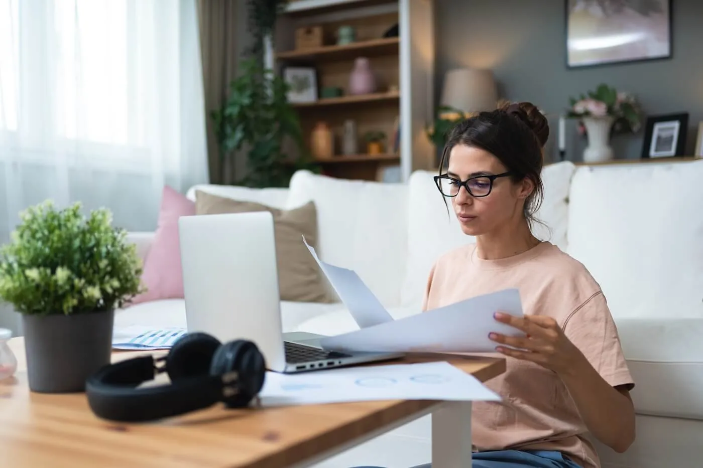 Focused young woman filing taxes at home while sitting on the floor next to a coffee table with an open laptop and reviewing the printed documents