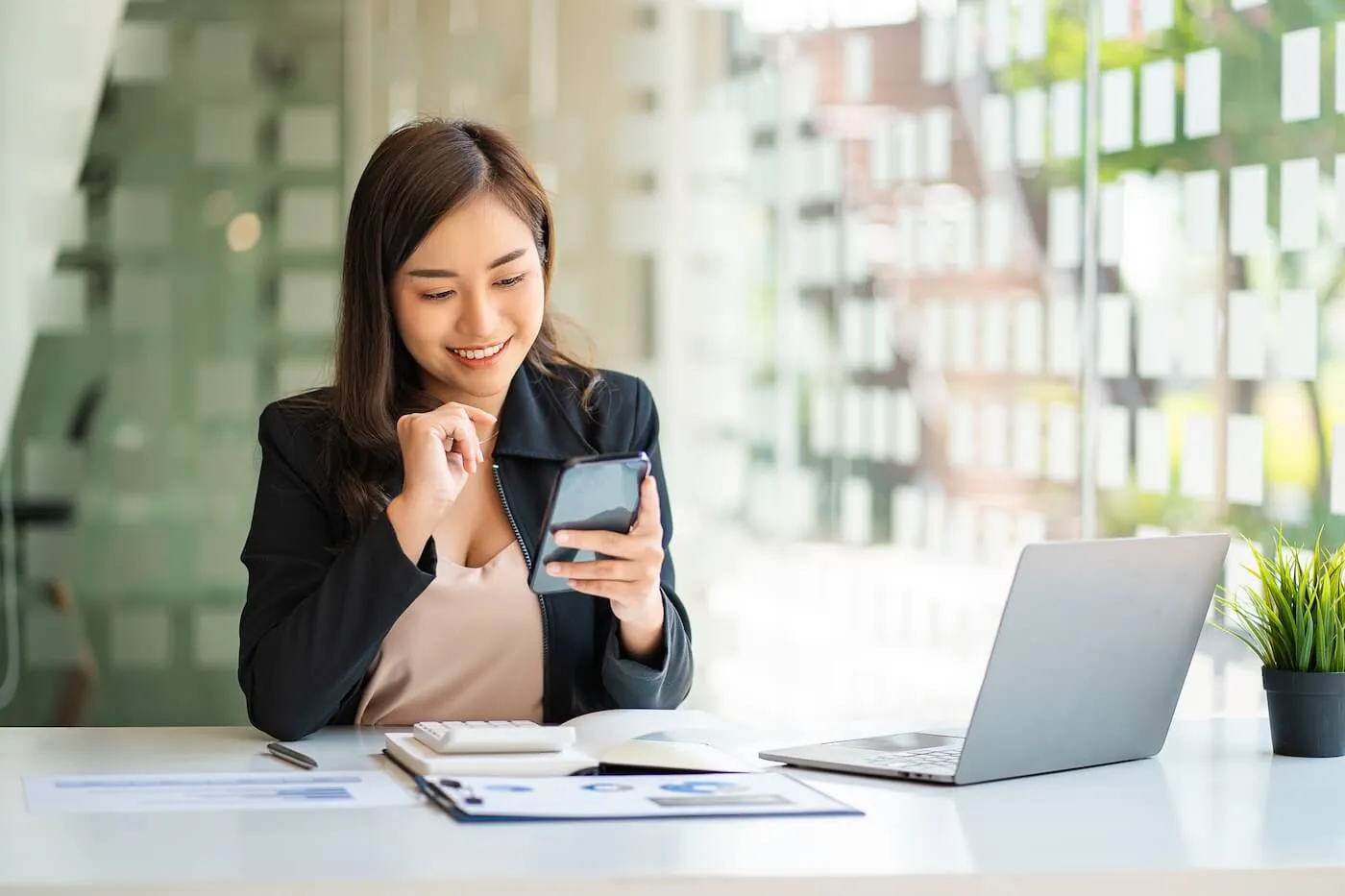 Smiling young woman in a business‑casual outfit checks her smartphone at her work desk, with an open laptop and documents beside her