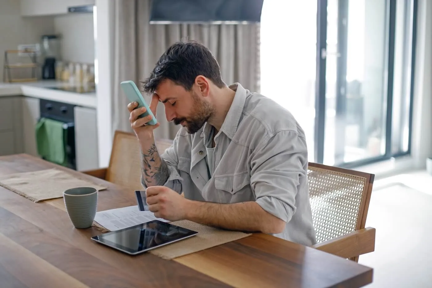 Concerned man sitting at a wooden dining table with eyes closed, holding a smartphone to the forehead and a credit card, with a printed document, tablet, and mug nearby; a kitchen is visible in the background.