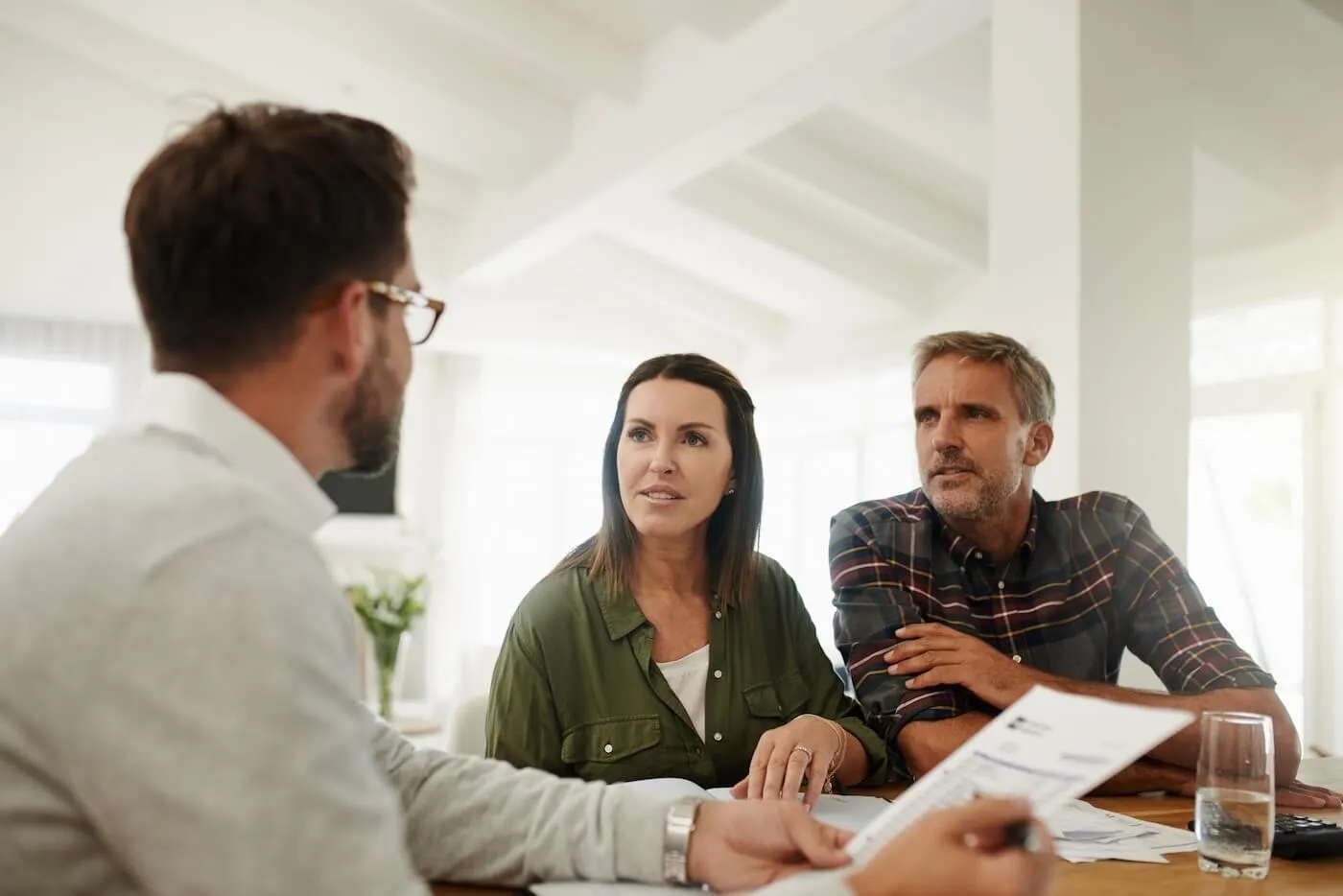 Focused couple sitting with a financial advisor, discussing documents, with papers and a calculator spread out in a bright home setting.