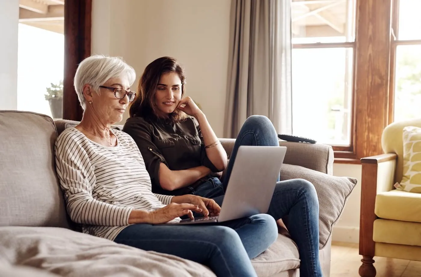 Adult daughter sitting on a sofa with her older mother, who is using a laptop
