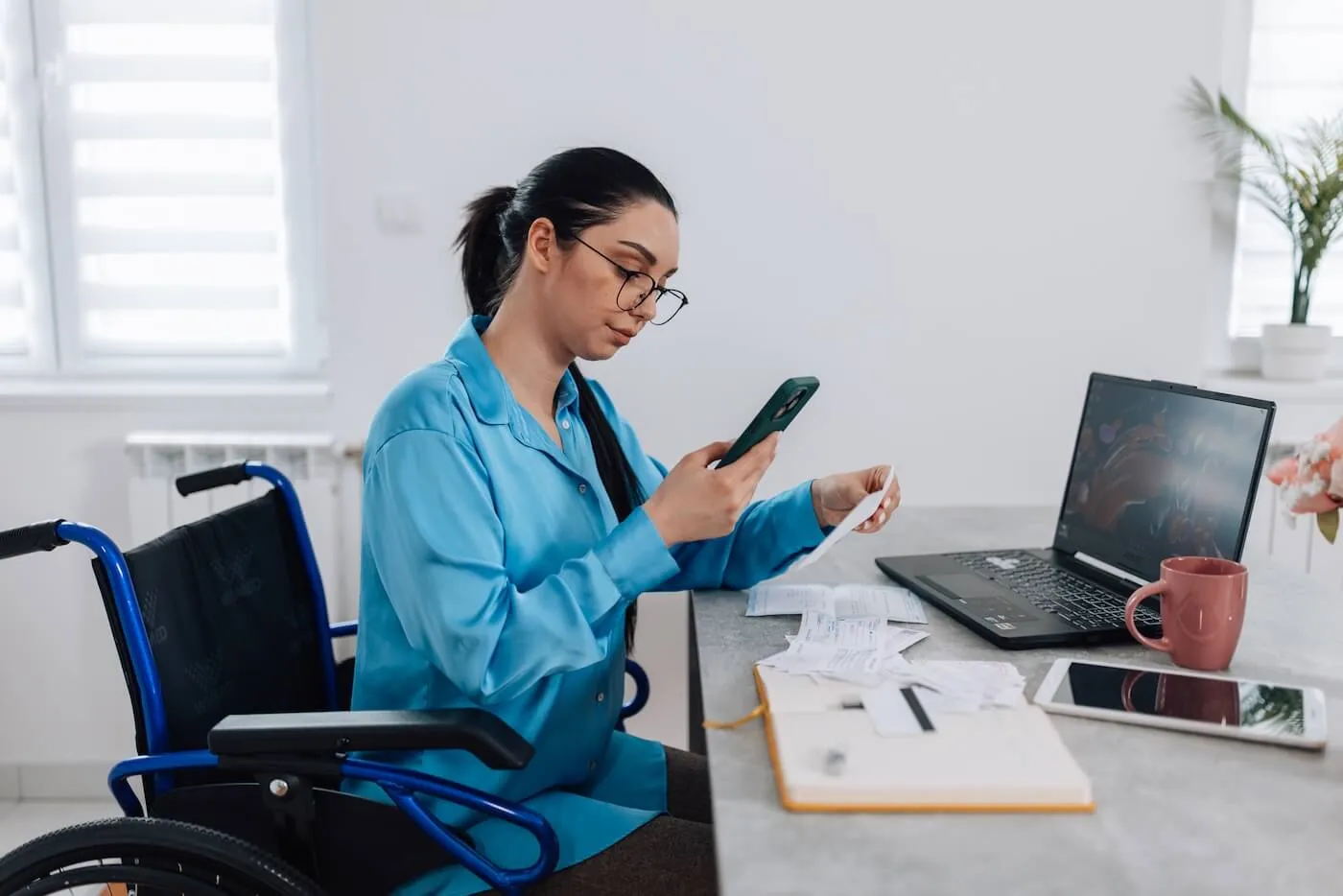 Focused young woman in a wheelchair sits at a desk reviewing receipts, holding a smartphone while checking a printed bill, with a laptop and documents spread out nearby.