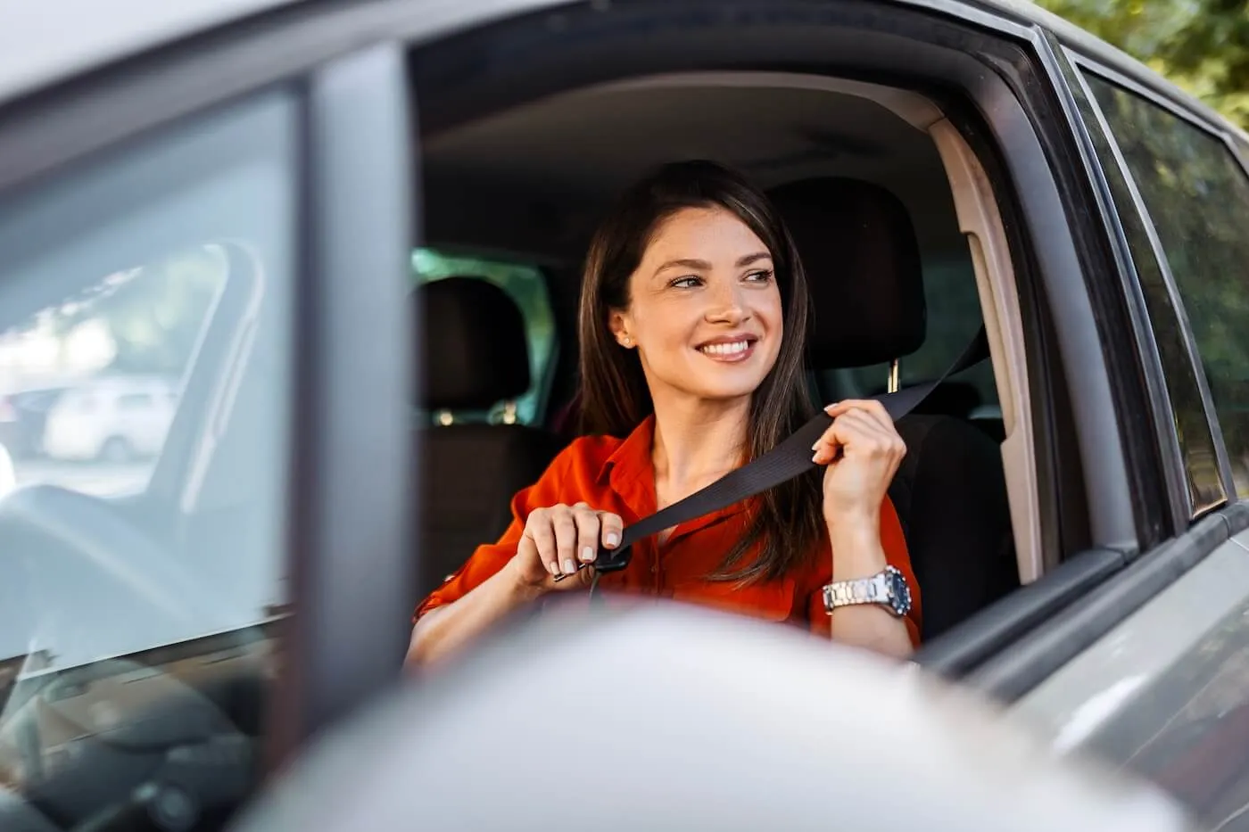 Smiling woman sits in the driver’s seat of a car and pulls a seat belt across the chest while preparing to buckle up