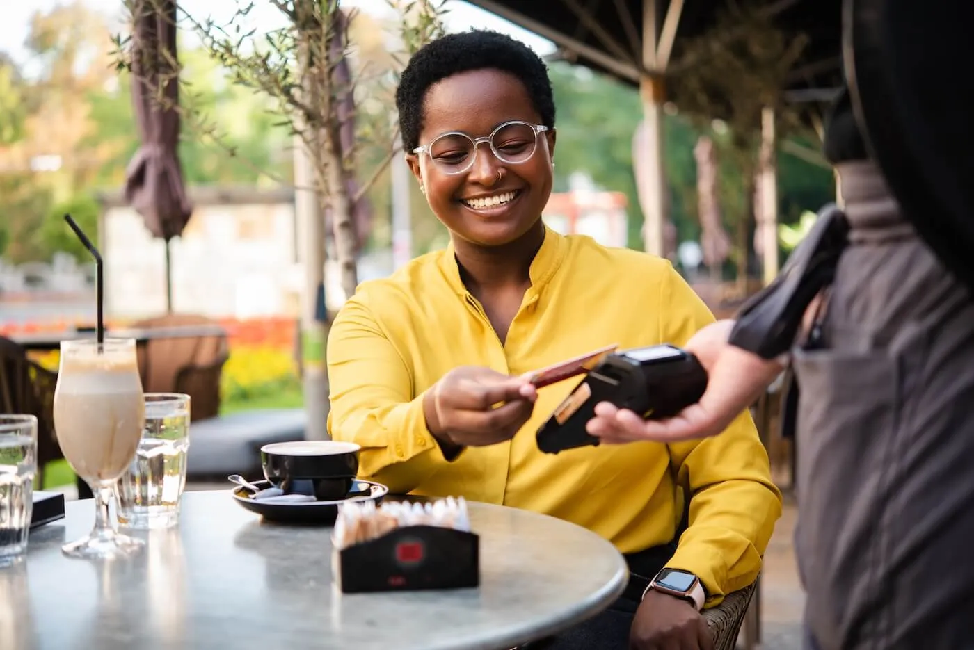 Smiling young woman seated at an outdoor café table hands a payment card to a waiter holding a portable card reader, with drinks, glassware, and a small container on the table, and trees, umbrellas, and café seating visible in the background.