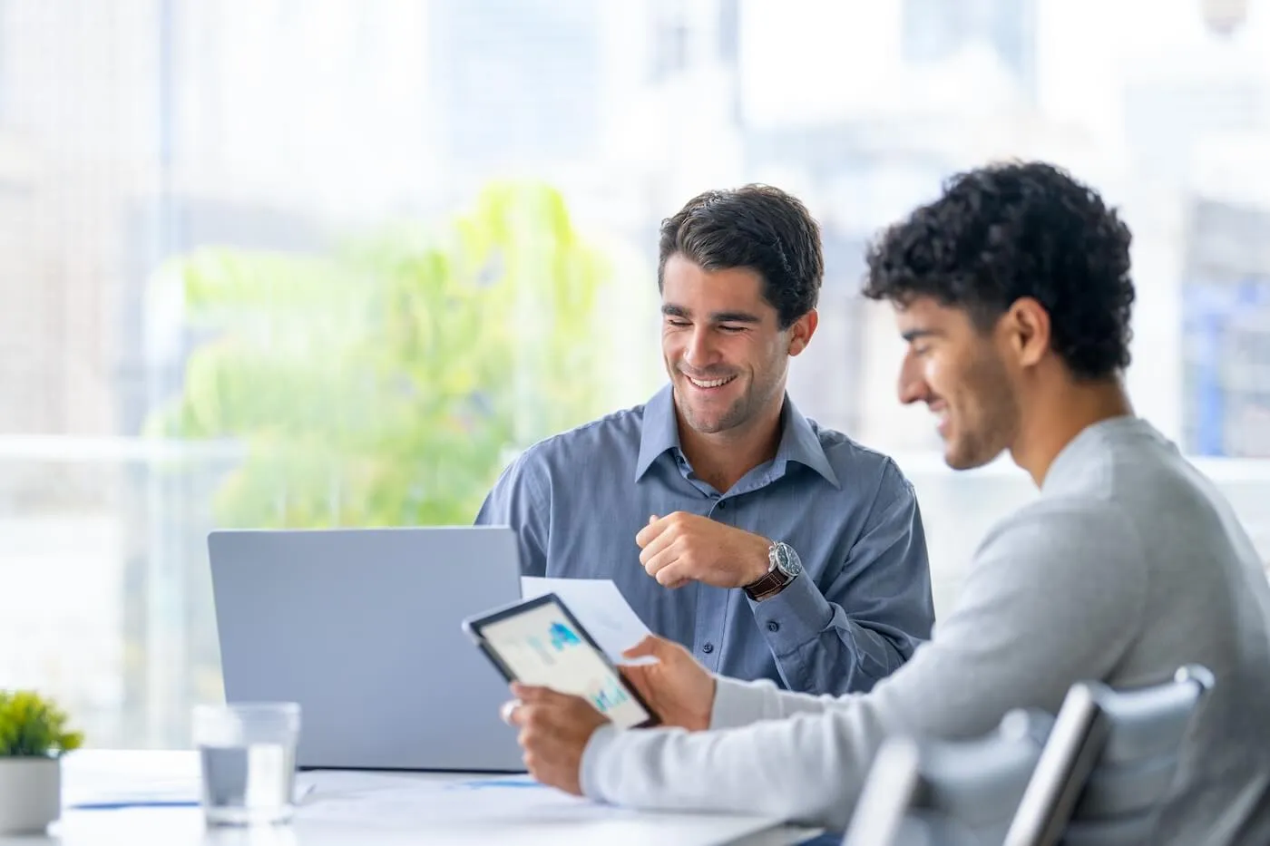 Two businessmen sit at a table reviewing data on a tablet and laptop together in a bright office.