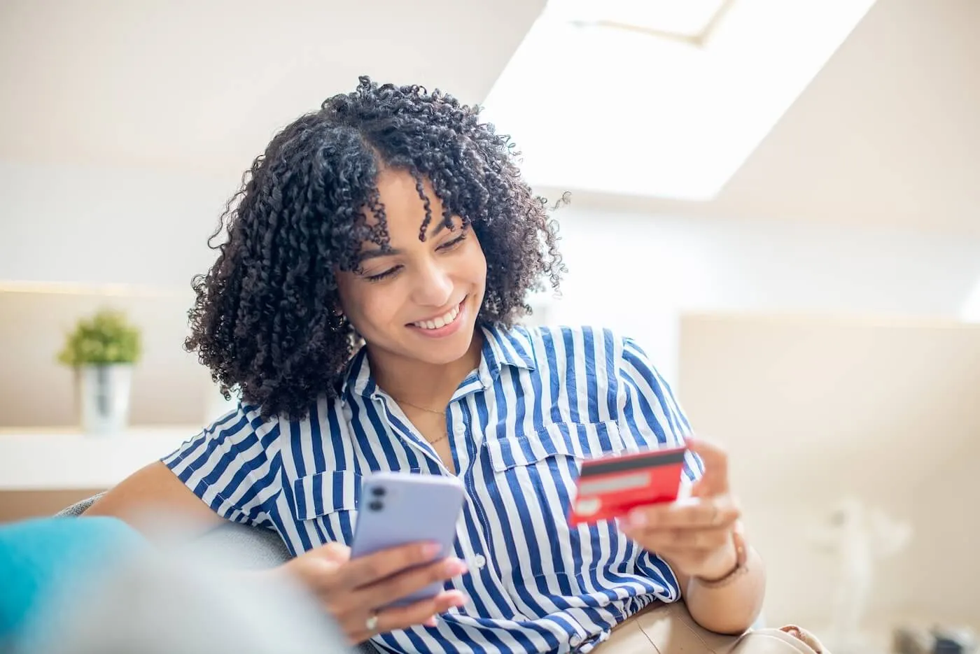 Smiling young woman sitting on a white couch and using a credit card on her smartphone