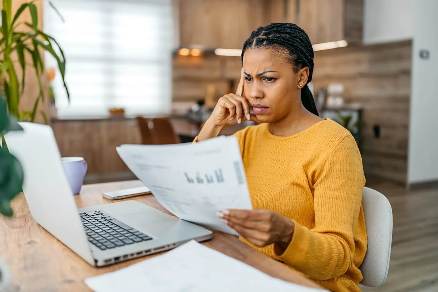 Concerned mature woman reviewing tax documents at a kitchen table, with a laptop open beside her