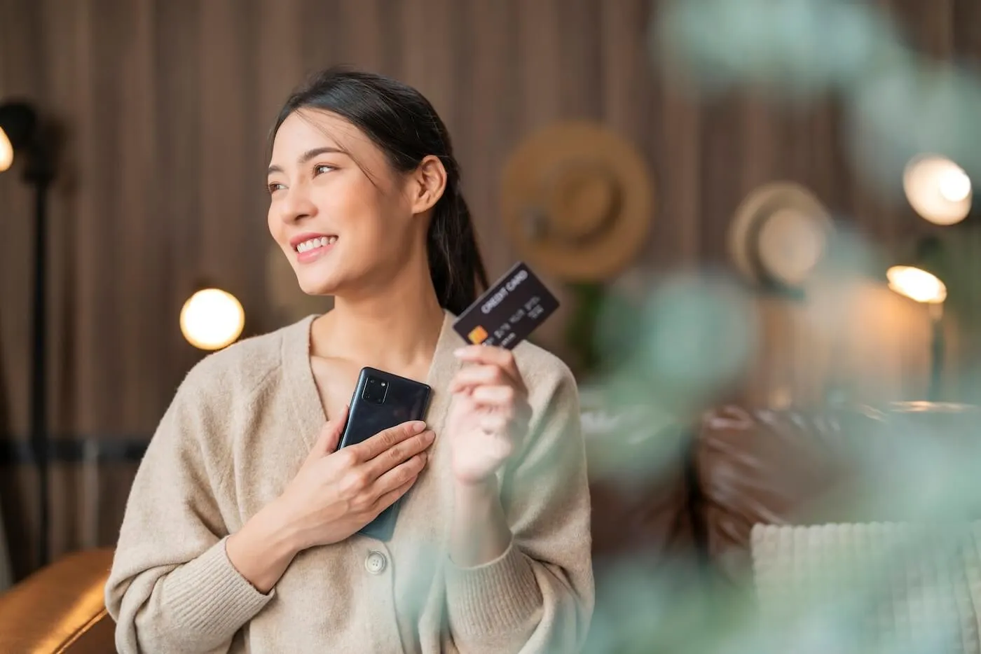 Happy young woman holding a credit card and smartphone while standing in a cozy living room