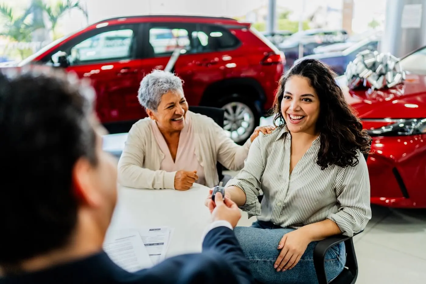 Three people seated at a desk inside a car dealership, with an agent handing a set of car keys across the table to a smiling young woman, while an older woman sits nearby; red vehicles, dealership lighting, and paperwork are visible in the background.