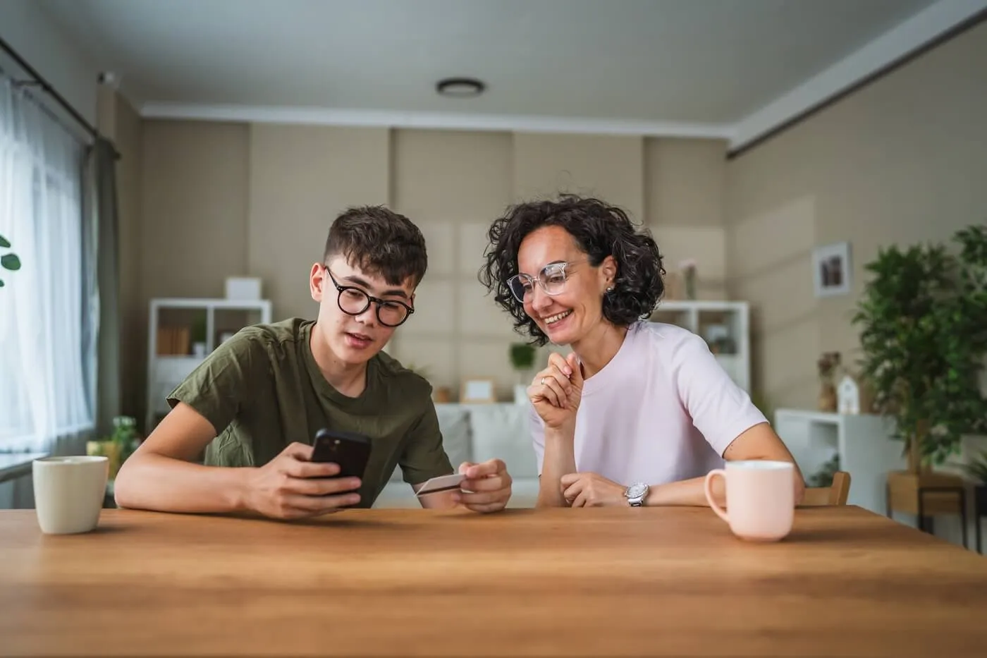 Mature woman and a teenage boy holding a credit card and looking at a smartphone together with mugs on the table