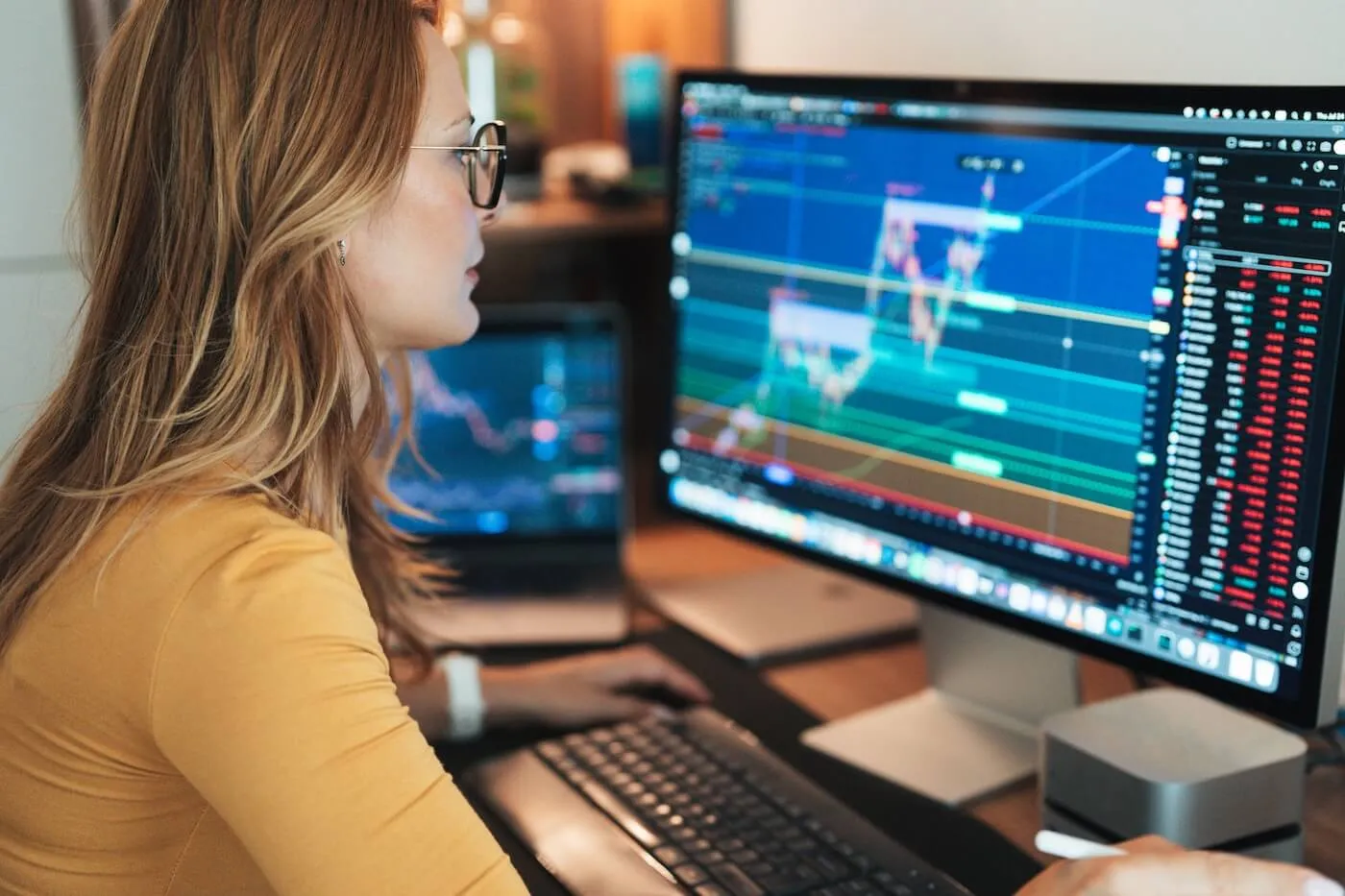 Focused woman working at a desktop computer displaying stock market charts and trading data, with multiple monitors showing financial graphs and market performance information.