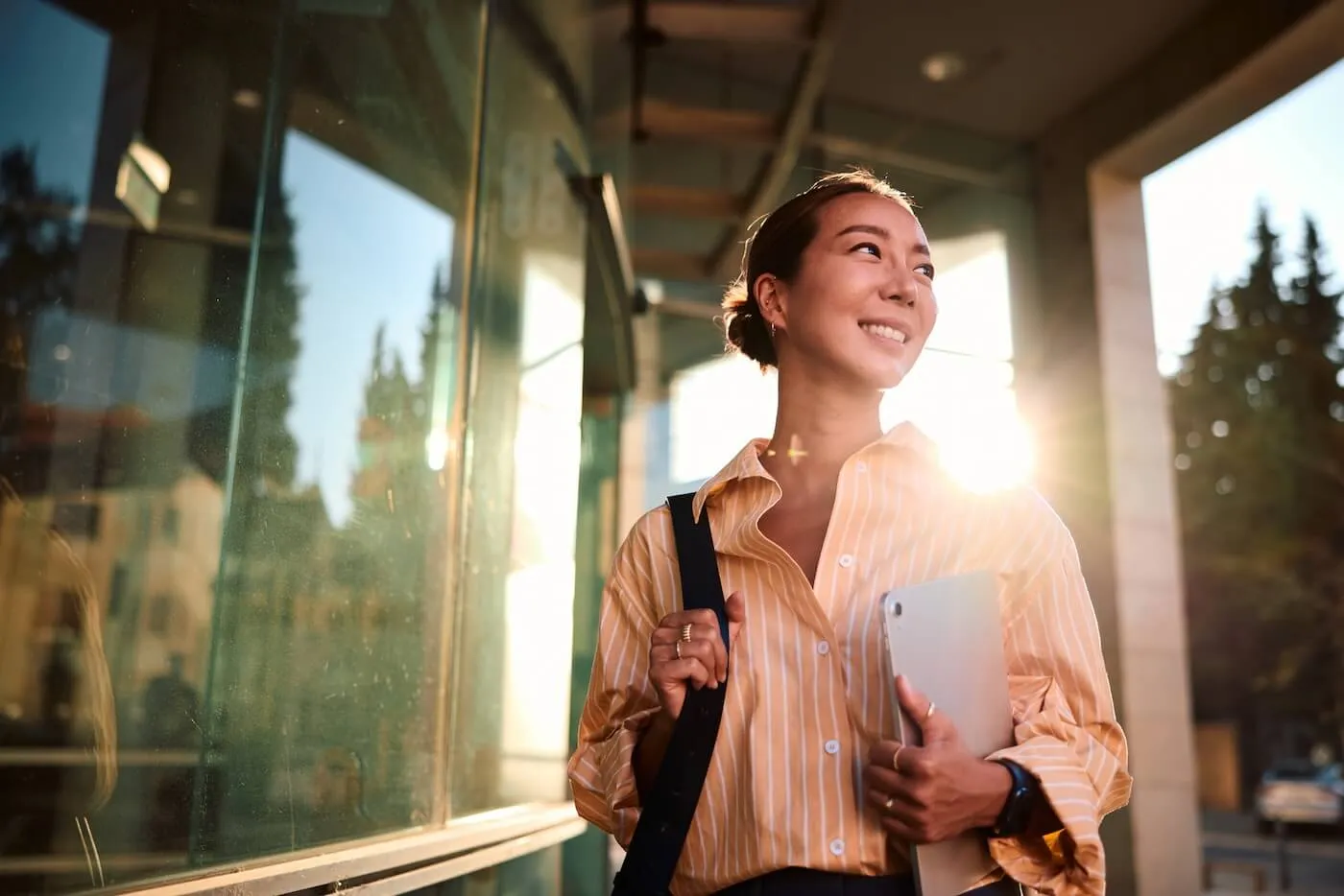 Smiling young woman walking outside a modern office building while carrying a tablet and shoulder bag, with sunlight reflecting off glass windows and trees visible in the background.