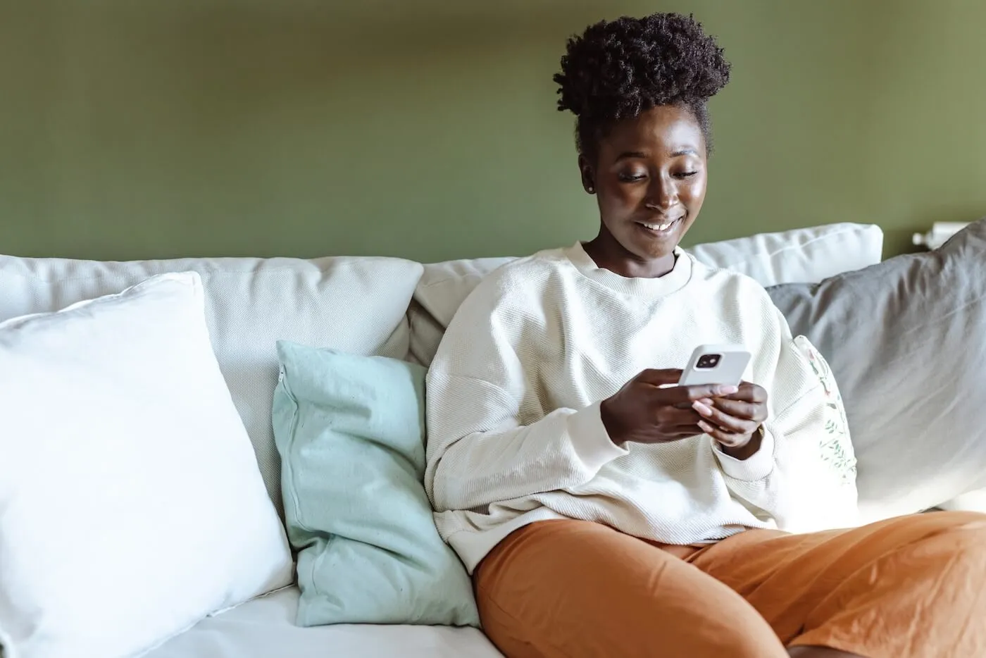 Smiling young woman sitting on a sofa in a bright living room while looking at a smartphone. Decorative pillows and a green accent wall are visible in the background.
