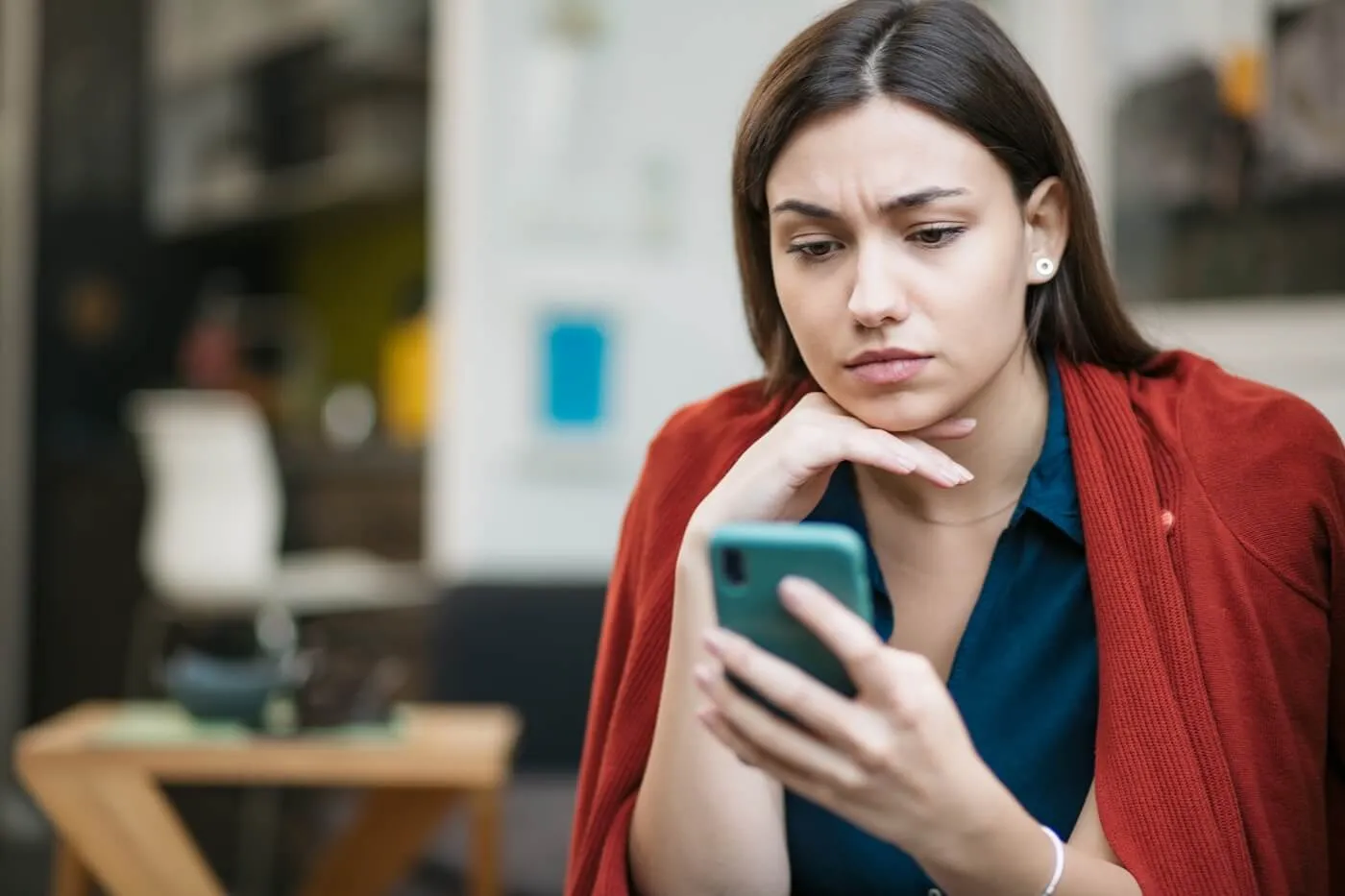 Concerned young woman seated at a café table looking at a smartphone, with one hand resting under the chin and a red cardigan draped over the shoulders. Blurred café seating and interior furnishings are visible in the background.