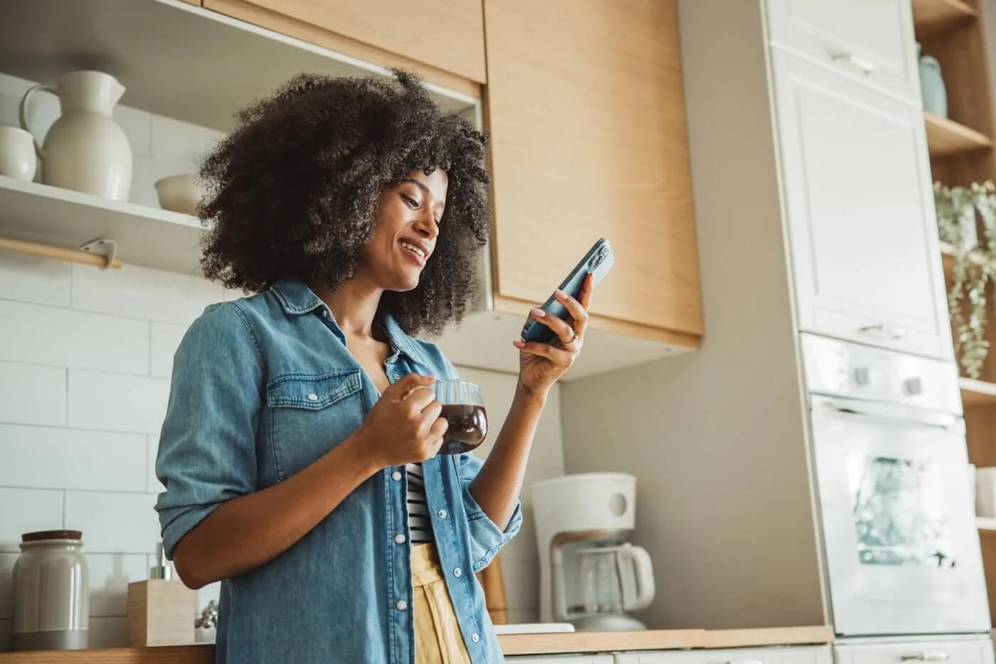 Smiling woman standing in a bright kitchen holding a mug of coffee and looking at a smartphone, with wooden cabinets, open shelving, and a coffee maker visible in the background.