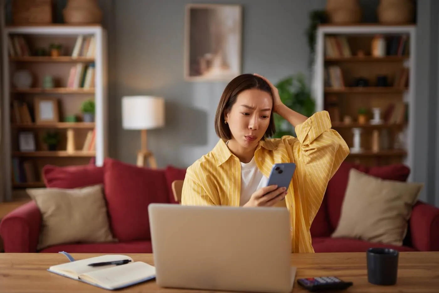 Concerned young woman sitting at a table in a living room, looking at a smartphone beside an open laptop, notebook, calculator, and coffee mug, with bookshelves and a sofa in the background.