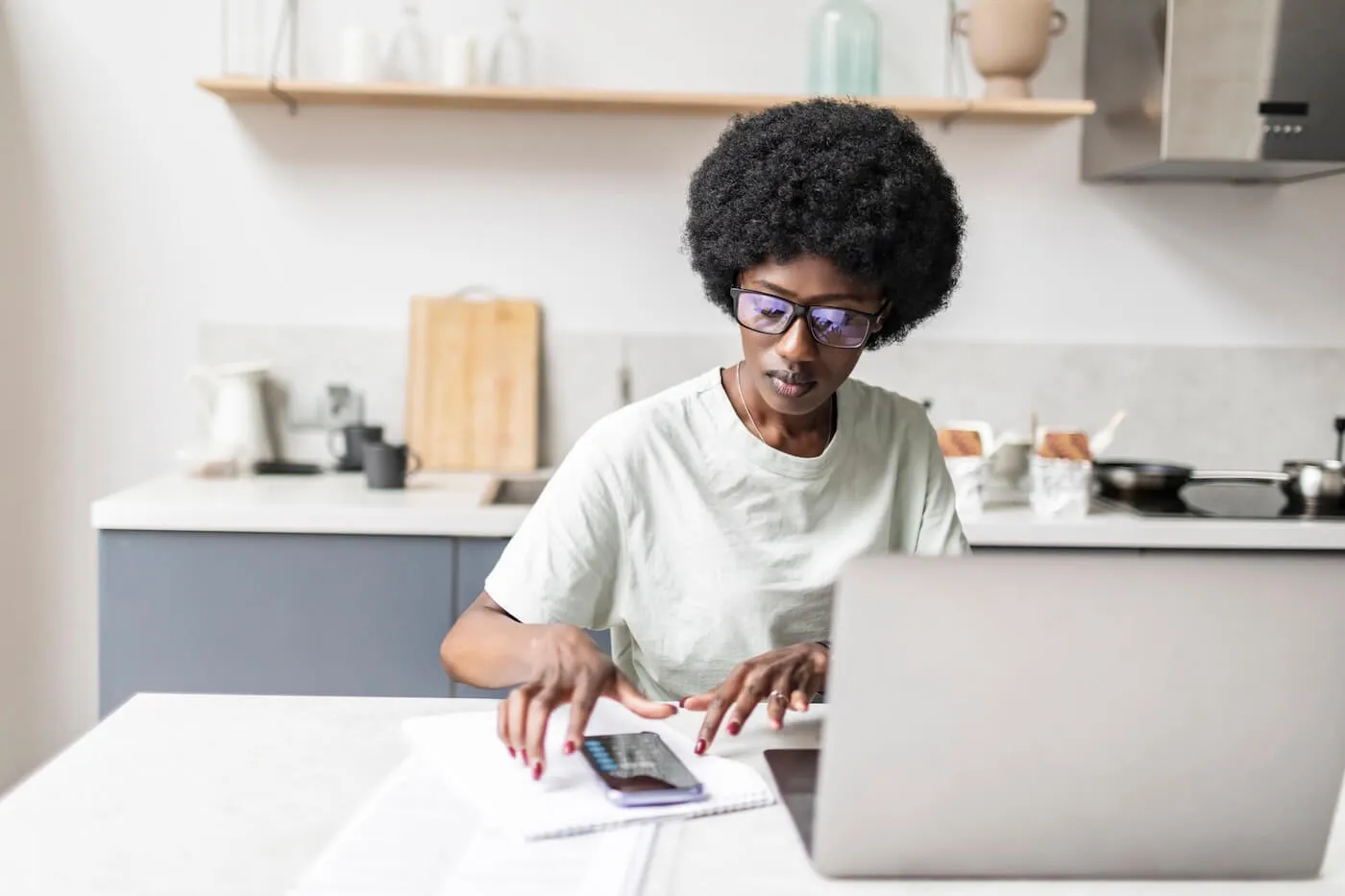 Focused woman seated at a kitchen table using a laptop and calculator while reviewing financial documents and notes, with household items and countertops visible in the background.