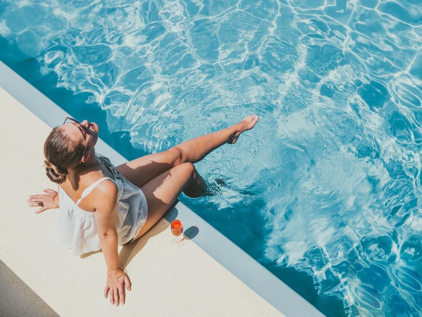 Woman in sunglasses sitting at the edge of a swimming pool with feet in the water, relaxing on a sunny day with a drink nearby.
