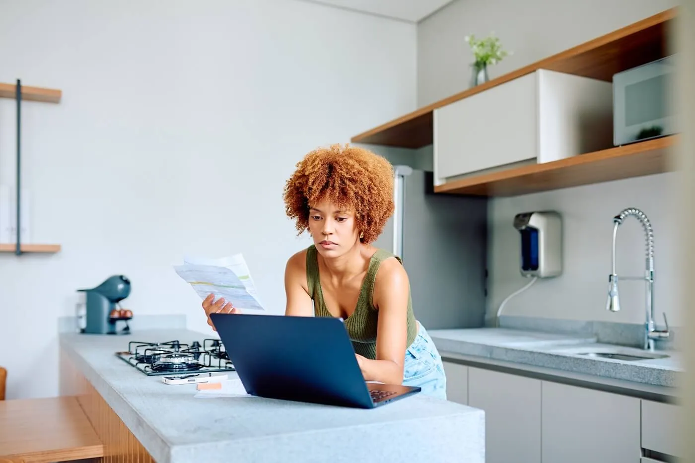 Focused young woman standing at a kitchen counter using a laptop while reviewing a printed document, with a smartphone and household items nearby in a modern home kitchen.