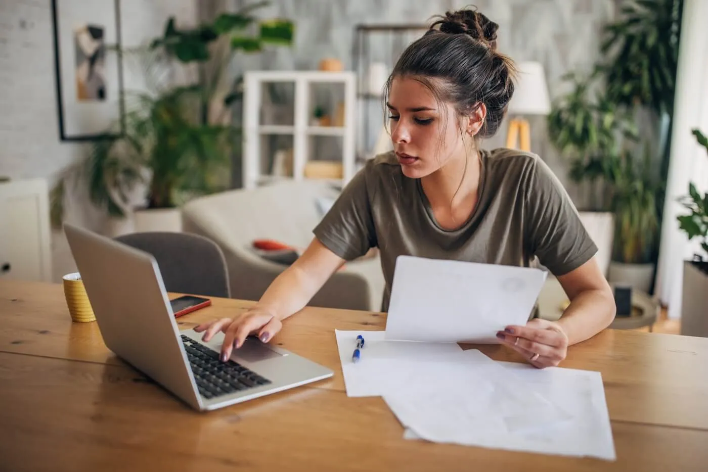 Focused woman seated at a wooden table in a home office, reviewing printed documents while using a laptop. Papers, a pen, a smartphone, and a coffee cup are arranged on the table, with houseplants and living room furniture visible in the background.