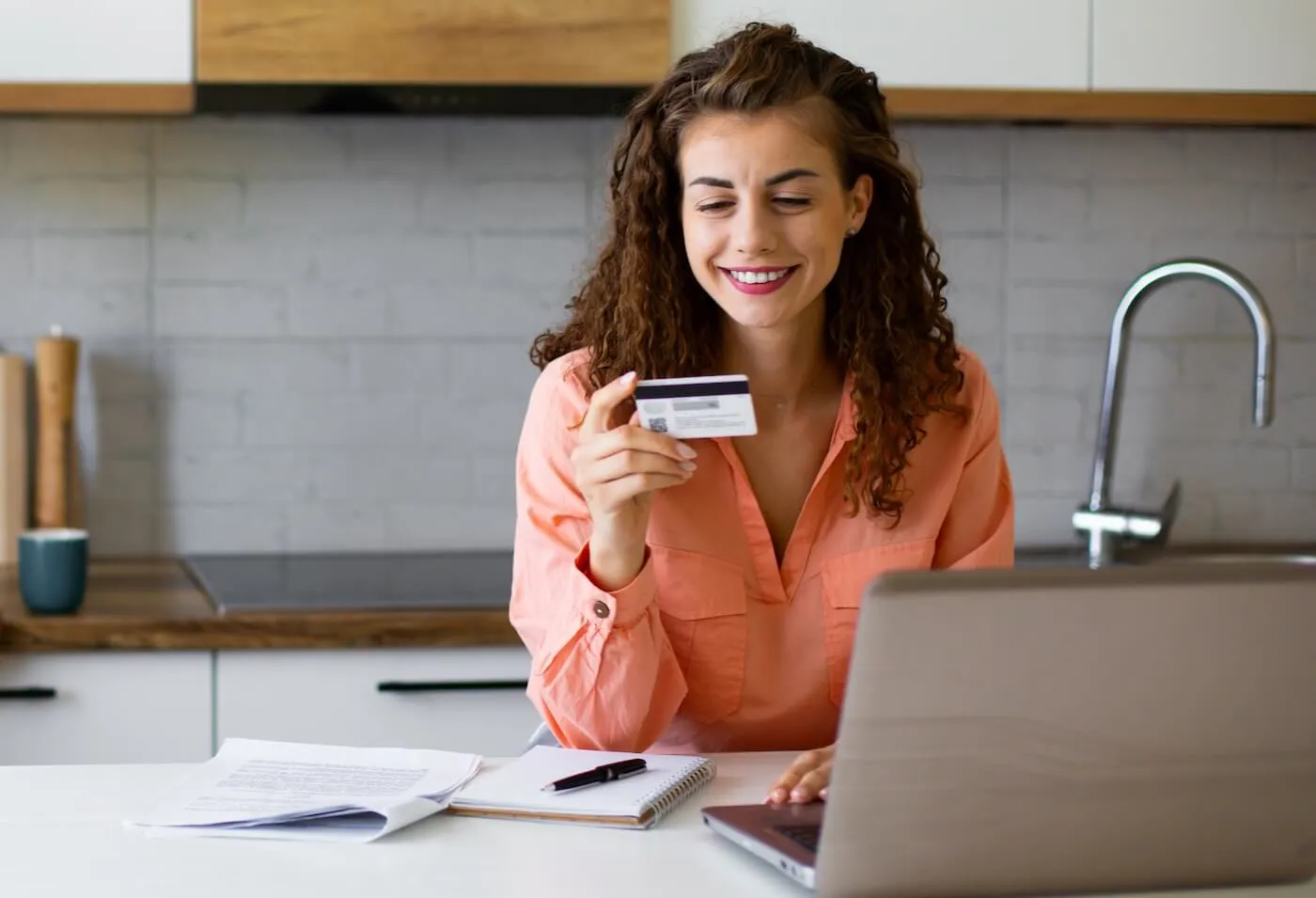 Smiling woman seated at a kitchen table holding a credit card while using a laptop, with a notebook and paperwork nearby.