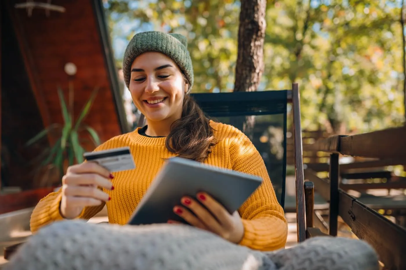 Smiling woman sitting on an outdoor deck holding a credit card and using a tablet, with a cabin-style building and trees in the background, illustrating online shopping or digital banking in a relaxed setting.