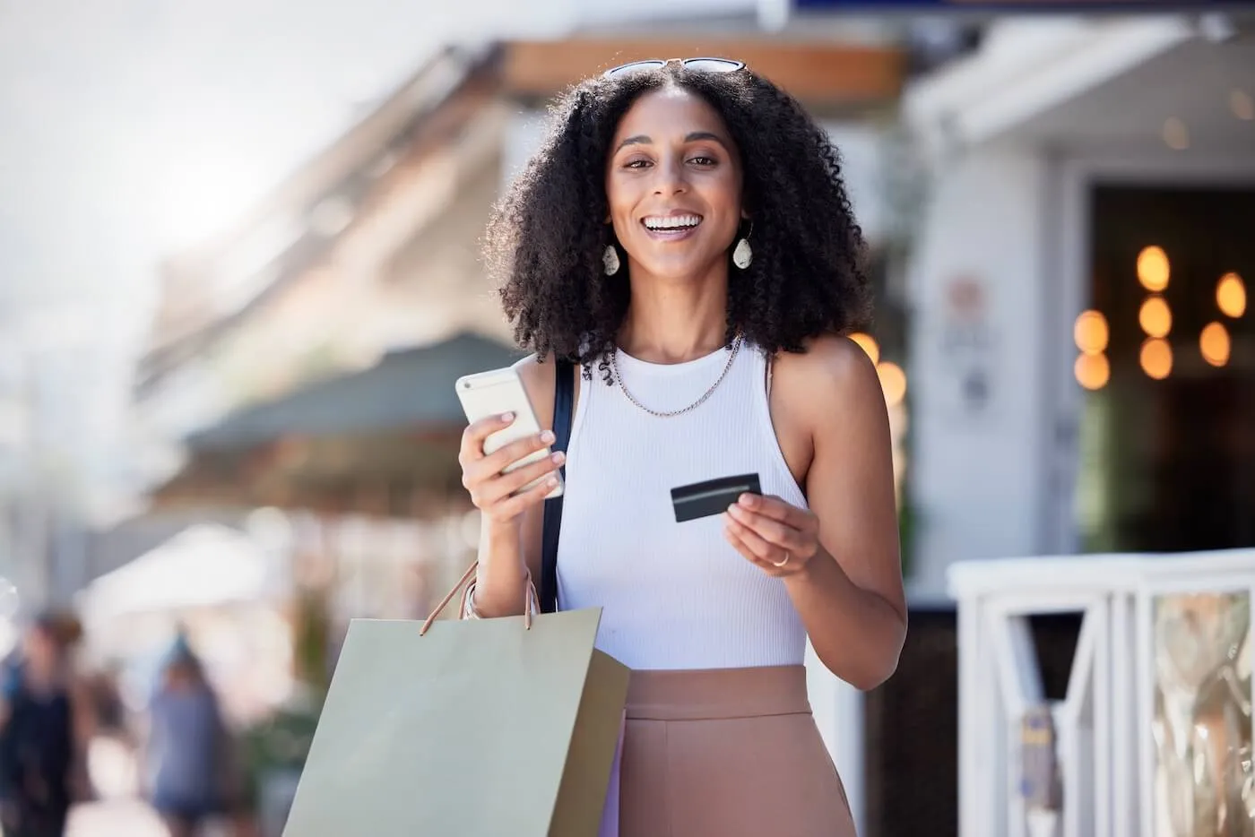 Happy woman standing outdoors holding a smartphone, payment card, and shopping bag, with storefronts and a pedestrian shopping area visible in the background.