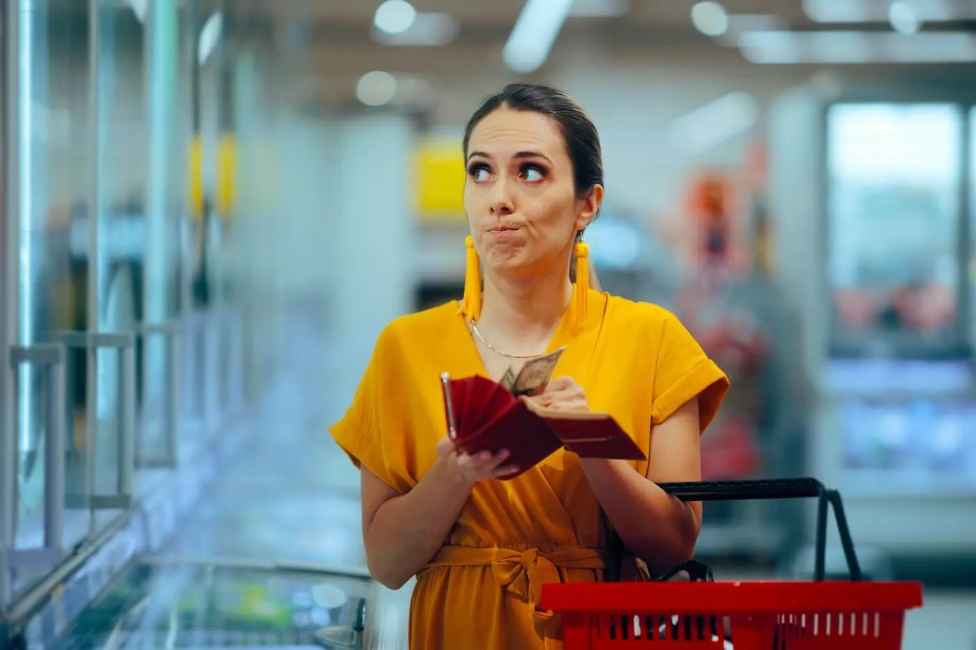 Concerned woman standing in a grocery store aisle holding an open wallet with cash visible and a red shopping basket on one arm, looking at refrigerated food shelves and comparing purchases to available funds.
