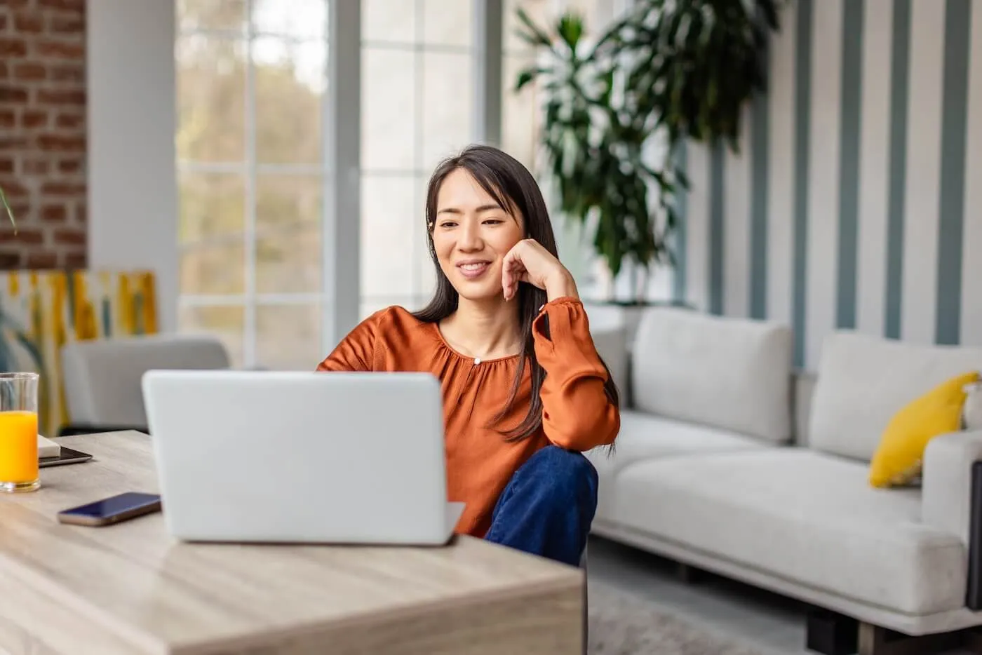 Smiling young woman seated at a table using a laptop in a bright living room, with a smartphone and a glass of orange juice on the table. Large windows, indoor plants, and a light-colored sofa are visible in the background.