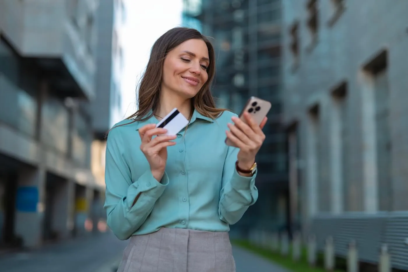 Smiling woman standing on a city street holding a credit card and using a smartphone, demonstrating mobile banking, digital payments, or online shopping.