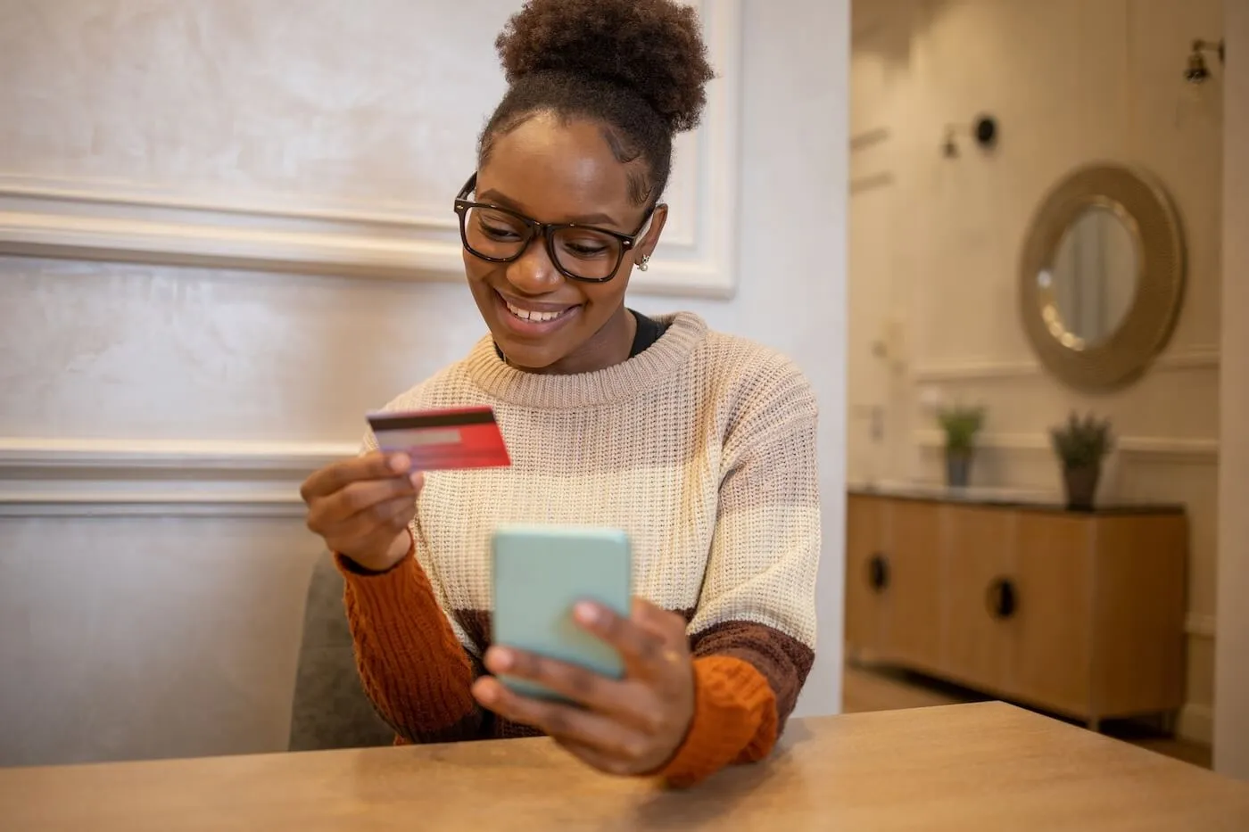 Smiling young woman sitting at a table using a smartphone while holding a credit card, illustrating online shopping, digital payments, mobile banking, or managing purchases from home. A decorative console table and wall mirror are visible in the background.