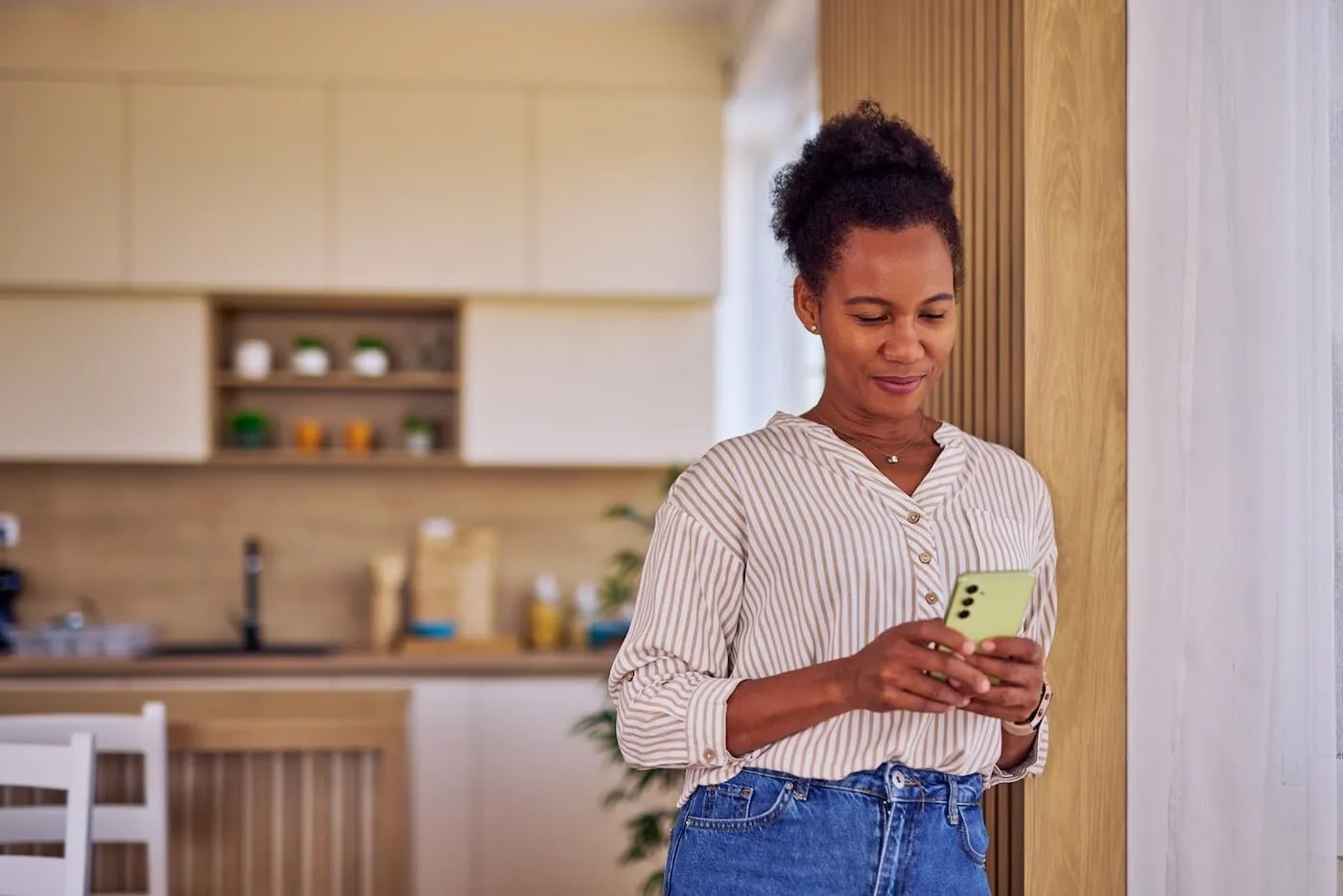 Smiling woman standing in a modern kitchen and using a smartphone near a window.
