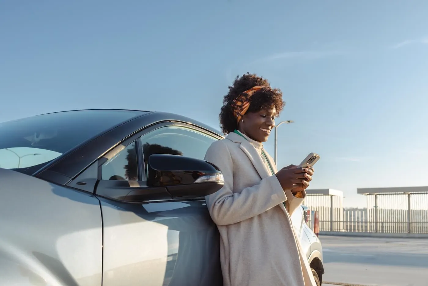 Smiling woman standing beside a parked vehicle and using a smartphone in an outdoor parking area, with clear skies and modern structures visible in the background.