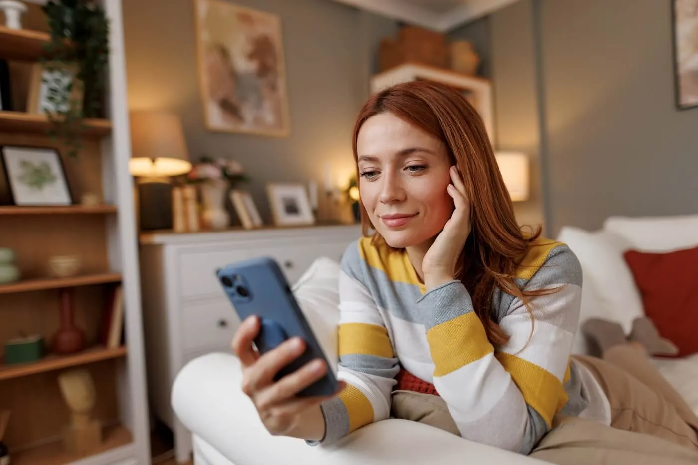Smiling woman reclining on a sofa in a cozy living room while looking at a smartphone. Bookshelves, framed artwork, decorative pillows, and soft lighting are visible in the background.