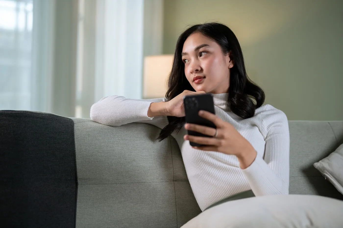 Pensive woman Person sitting on a sofa in a living room, holding a smartphone while resting an arm on the back of the couch, with soft indoor lighting and a floor lamp visible in the background.