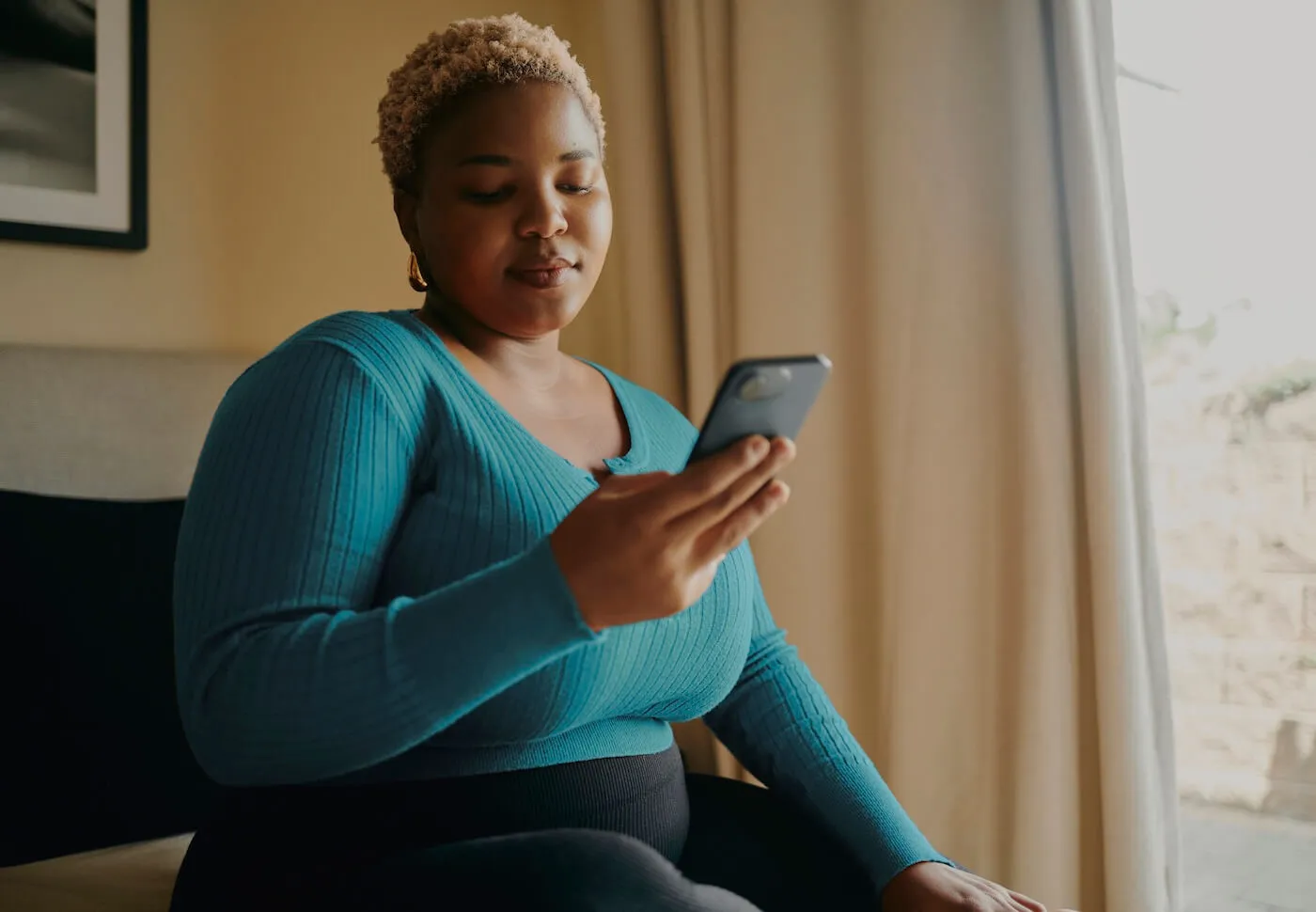 Focused woman seated in an armchair near a window, looking at a smartphone, wearing a blue sweater in a softly lit indoor setting with curtains and framed wall art in the background.