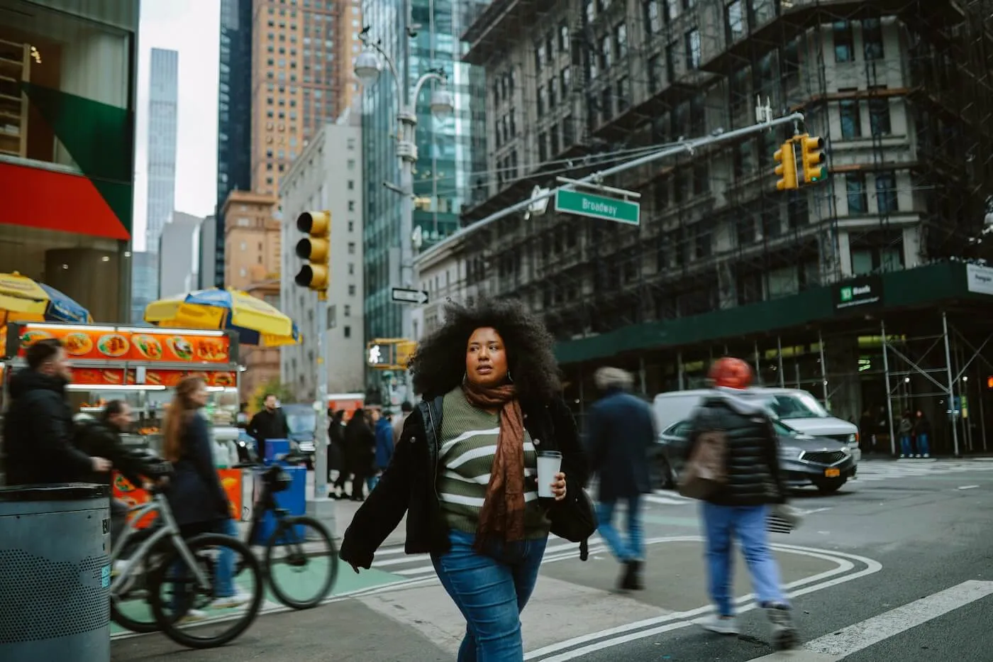 Focused woman carrying a takeaway coffee cup while walking through the NYC intersection surrounded by pedestrians, traffic, food carts, and tall office buildings. Crosswalk markings, street signs, and urban storefronts are visible in the downtown setting.