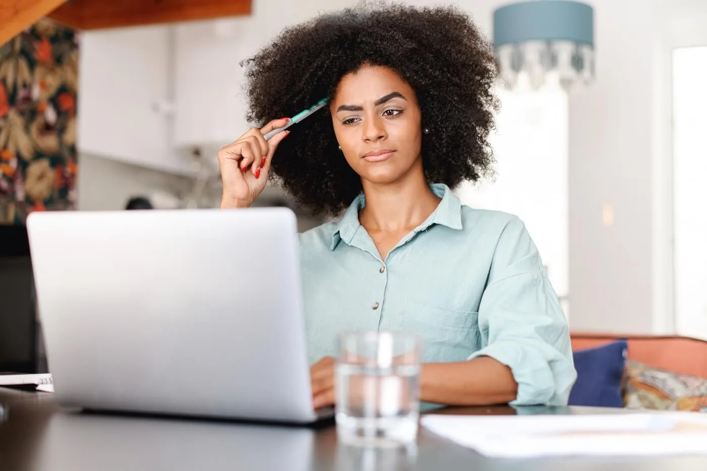 Focused young woman seated at a table using a laptop in a home setting, holding a pen near the forehead while reviewing information on the computer screen. A glass of water and documents are on the table, with a living area visible in the background.